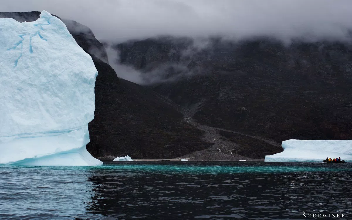 eisberg im wasser von einem winzigen schlauchboot begleitet als verdeutlichung des grössen-kontrasts