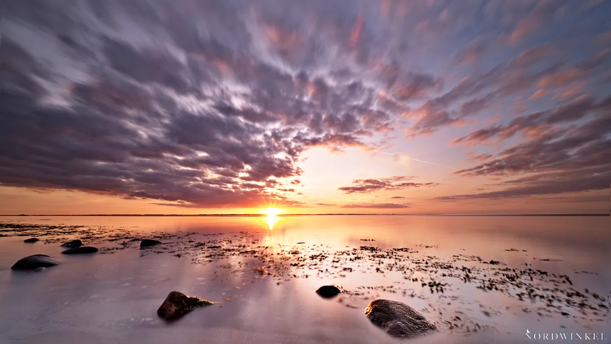 sonnenuntergang am strand mit steinen und leuchtend roten wolken als motivation fürs fotografieren lernen