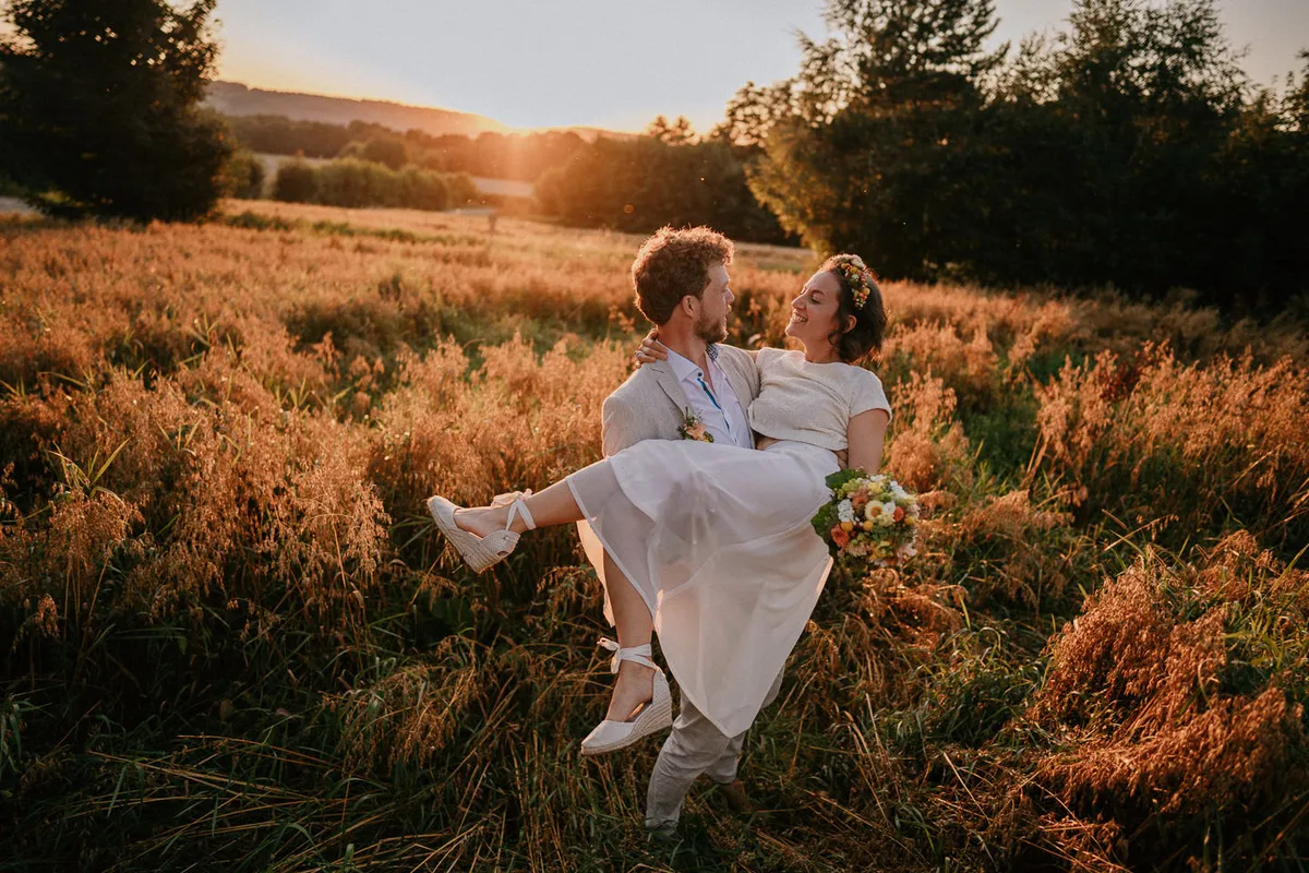 bräutigam trägt braut auf händen durch eine wildwiese im sonnenuntergang