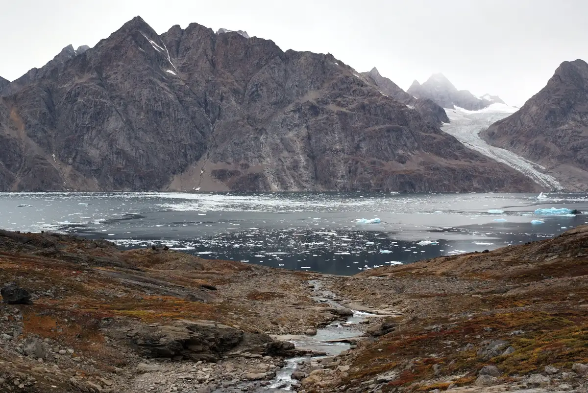 fluss fließt in eis bedeckten fjord mit steil aufragenden gipfeln und einem gletscher im hintergrund