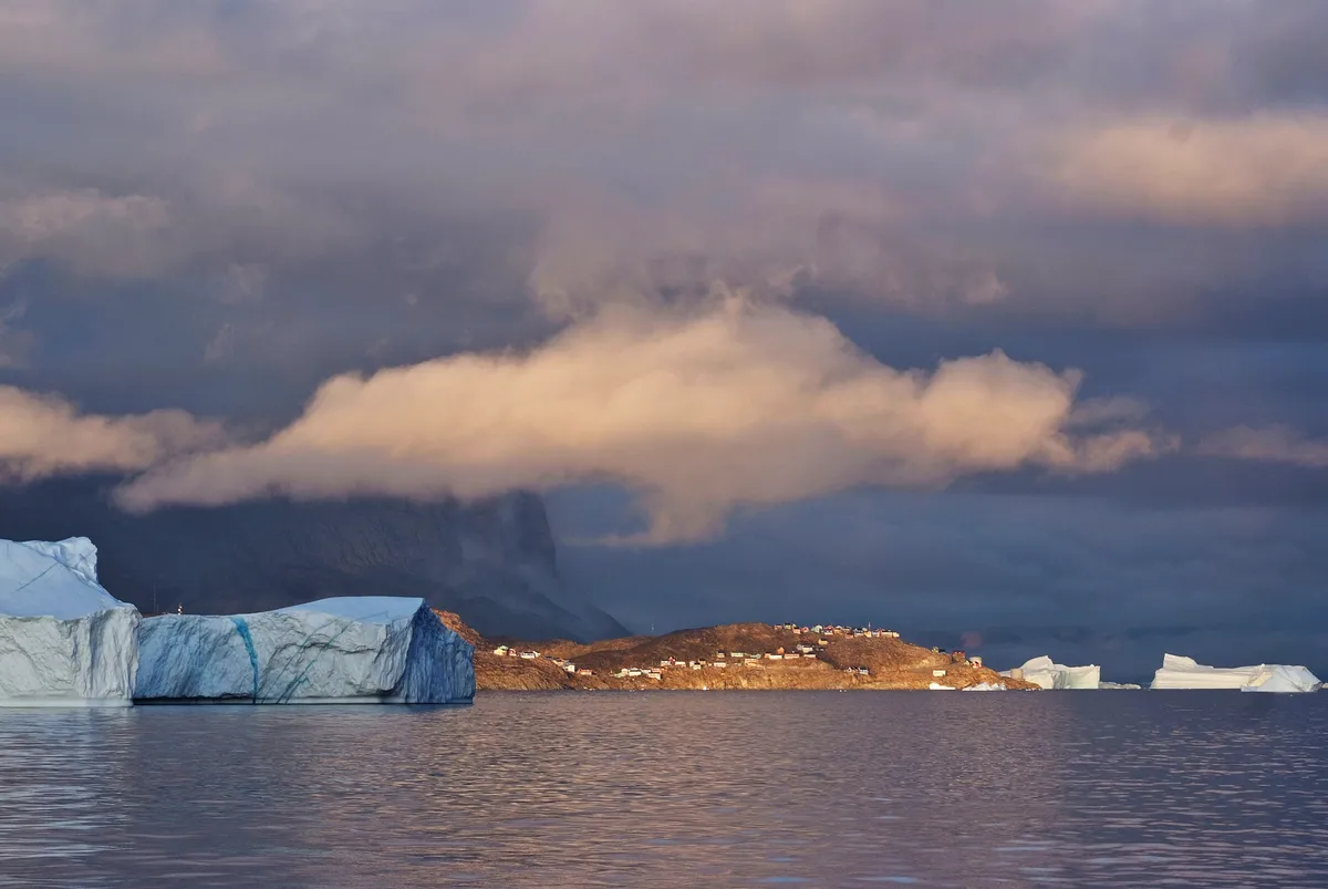 stadt uummannaq am abend hinter eisbergen und tiefhängenden angestrahlten wolken zum sonnenuntergang